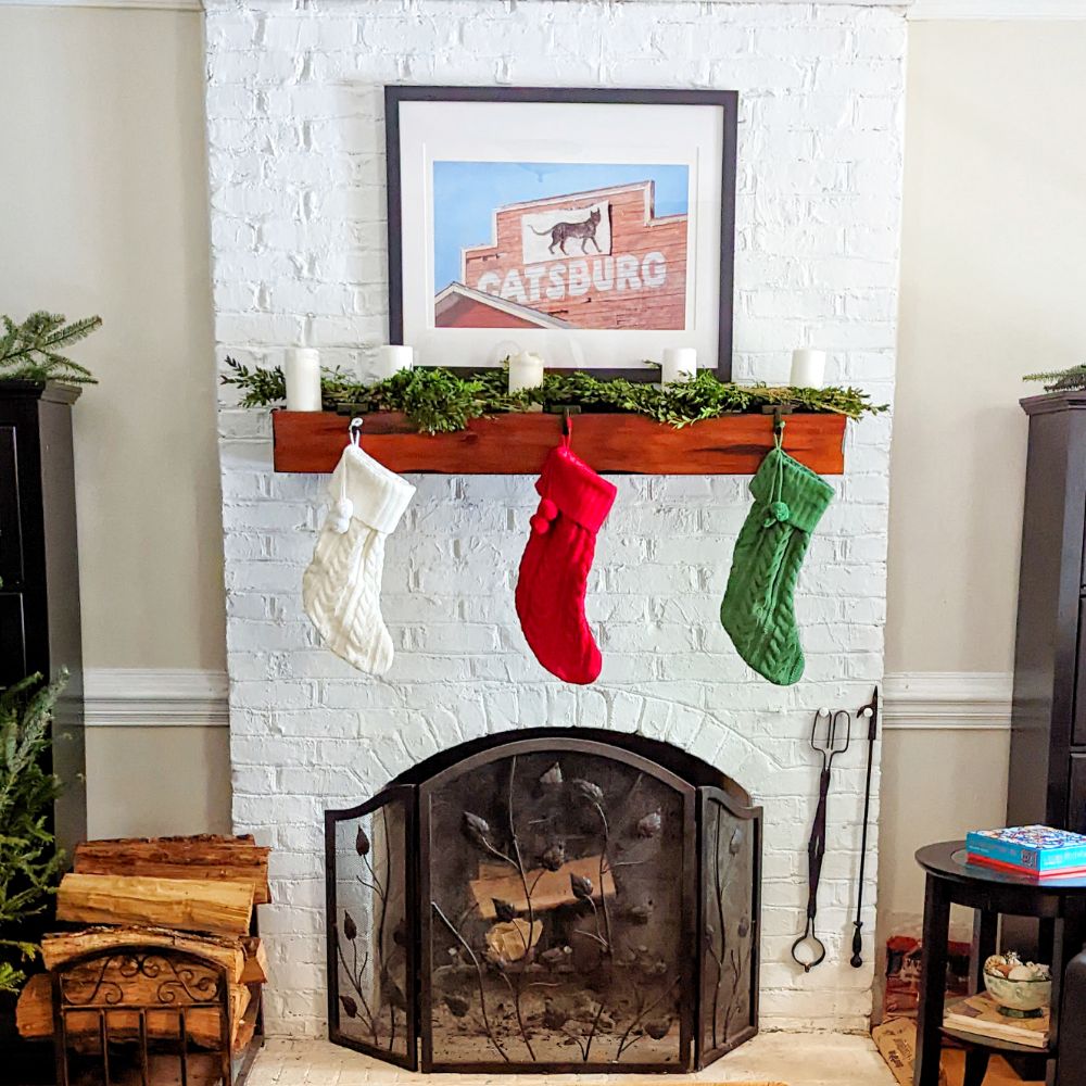 A brick fireplace painted white, a wide dark mantel hangs on the fireplace. Cream, red, and green Christmas stockings hang from the mantel. Greenery sits on the mantel with white candles spread through it. Above all this hangs a print of a painting of a red wooden building with a sign reading 'Catsburg' and a silhouette of a cat 