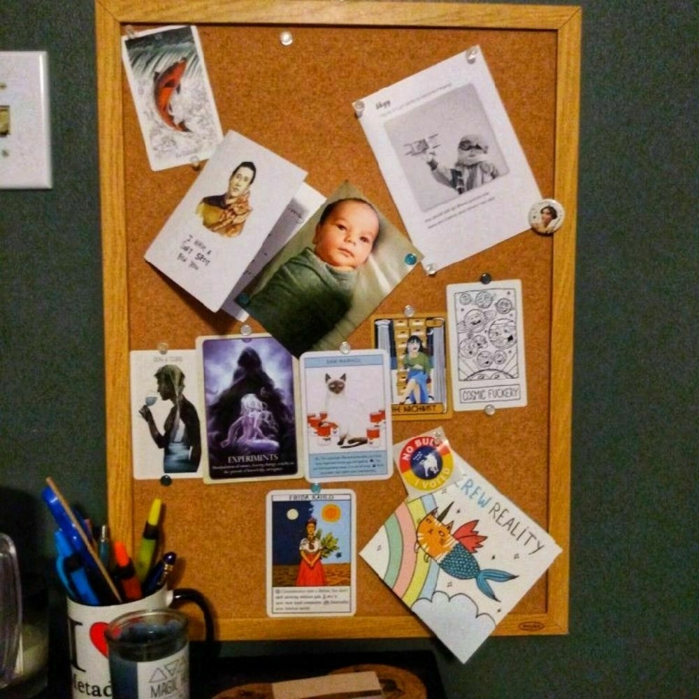 A cork board with several items pinned to it. Most of the items are tarot or oracle cards. There are also a photo of a swaddled baby and 3 greeting cards, as well as an "I voted" sticker. 