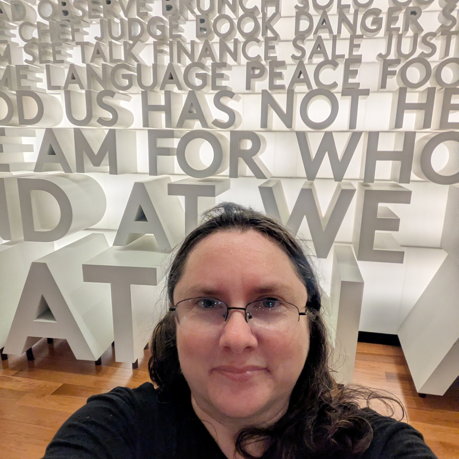 A white woman with dark hair and glasses stands in front of a wall with several words in 3d on it. The word 'BOOK' is centered over her head.